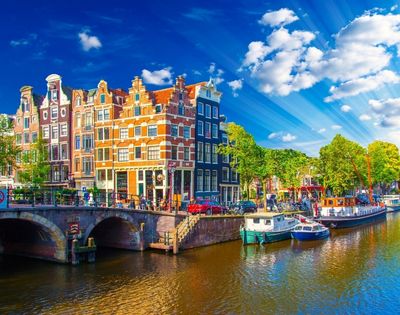 Traditional canal houses and boats along a canal in Amsterdam under a bright blue sky with sunbeams.