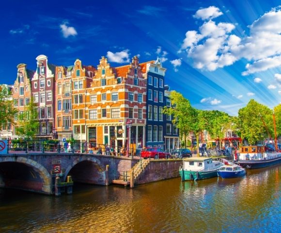 Traditional canal houses and boats along a canal in Amsterdam under a bright blue sky with sunbeams.