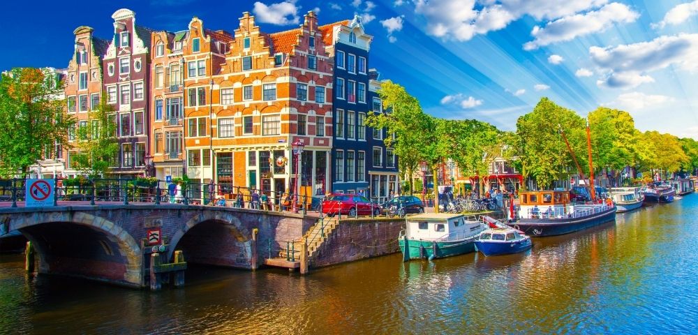 Traditional canal houses and boats along a canal in Amsterdam under a bright blue sky with sunbeams.
