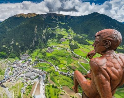 A bronze statue of a pensive male figure sits overlooking a valley town nestled between lush green mountains.