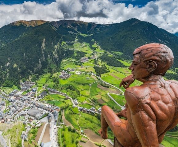 A bronze statue of a pensive male figure sits overlooking a valley town nestled between lush green mountains.