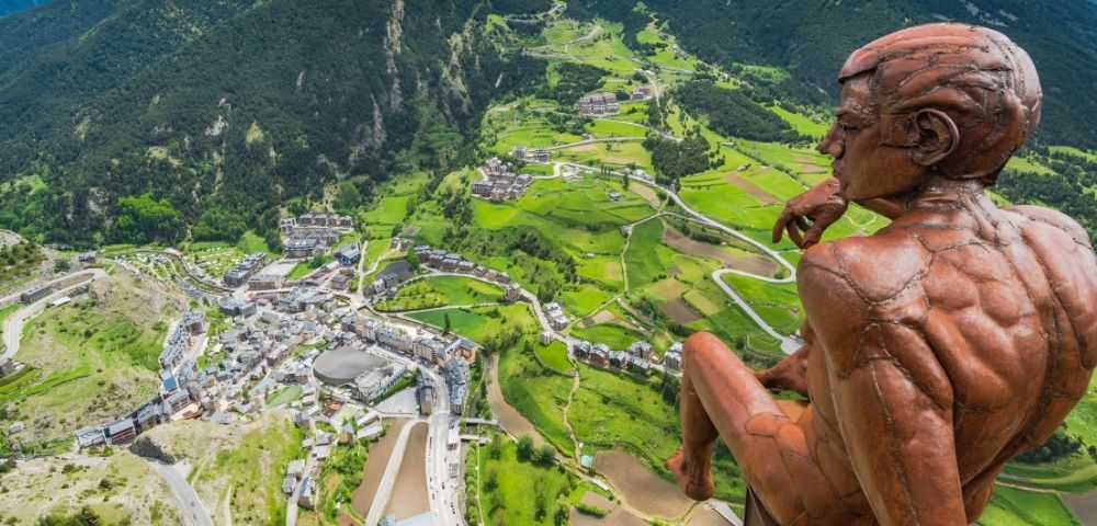 A bronze statue of a pensive male figure sits overlooking a valley town nestled between lush green mountains.