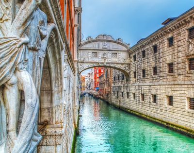 A statue on the left overlooks a turquoise canal in Venice, with the Bridge of Sighs arching over it.