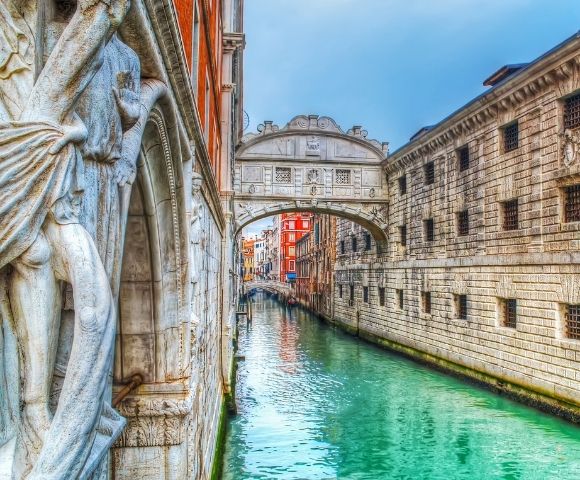 A statue on the left overlooks a turquoise canal in Venice, with the Bridge of Sighs arching over it.