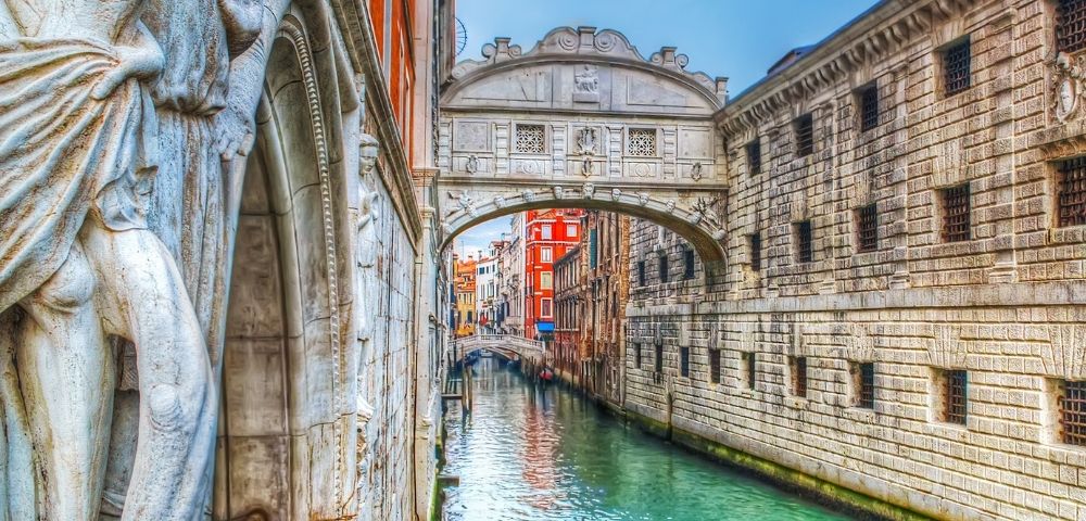 A statue on the left overlooks a turquoise canal in Venice, with the Bridge of Sighs arching over it.