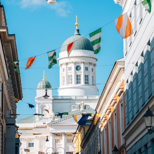 A sunny street scene with colorful flags strung between buildings, leading to a grand white cathedral with green domes under a bright blue sky.