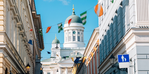A sunny street scene with colorful flags strung between buildings, leading to a grand white cathedral with green domes under a bright blue sky.