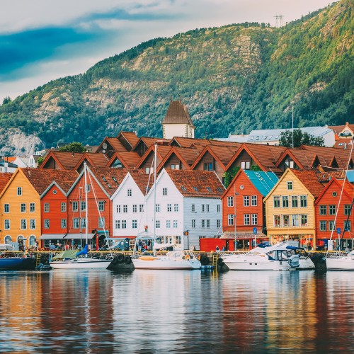 Colorful wooden houses with red roofs line a waterfront in front of a forested mountain. Boats are docked, reflecting a serene, picturesque setting.