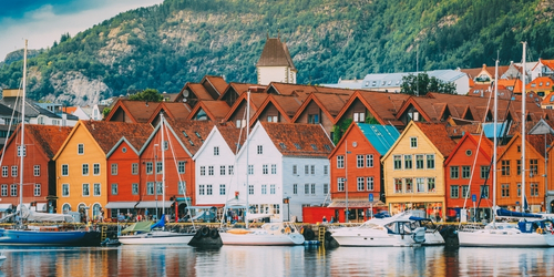 Colorful wooden houses with red roofs line a waterfront in front of a forested mountain. Boats are docked, reflecting a serene, picturesque setting.