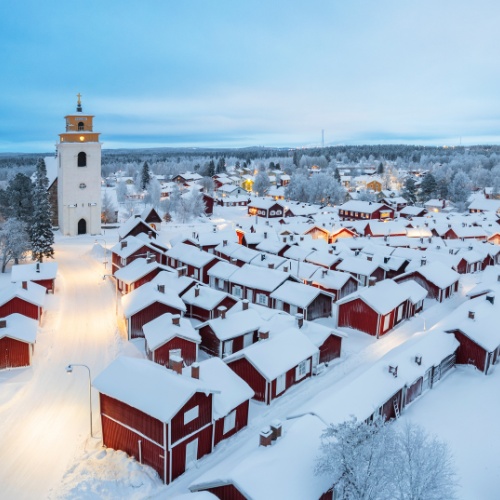Snow-covered village with red wooden houses and a tall church tower. Soft, warm streetlights create a serene, wintery evening atmosphere.