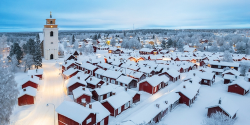 Snow-covered village with red wooden houses and a tall church tower. Soft, warm streetlights create a serene, wintery evening atmosphere.