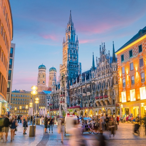 A bustling evening scene at Munich's Marienplatz, featuring the illuminated New Town Hall, Frauenkirche in the background, and blurred pedestrians.