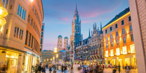 A bustling evening scene at Munich's Marienplatz, featuring the illuminated New Town Hall, Frauenkirche in the background, and blurred pedestrians.
