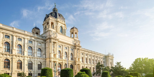 Grand, historic building in Vienna with ornate architecture and large dome, surrounded by manicured gardens under a clear blue sky. Majestic and serene.