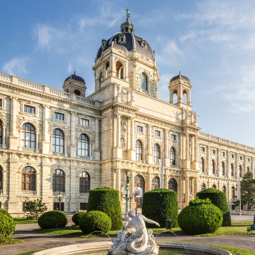 Grand, historic building in Vienna with ornate architecture and large dome, surrounded by manicured gardens under a clear blue sky. Majestic and serene.