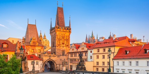 Charles Bridge in Prague at sunrise, featuring Gothic towers and statues. Warm light illuminates red-roofed buildings, creating a serene atmosphere.