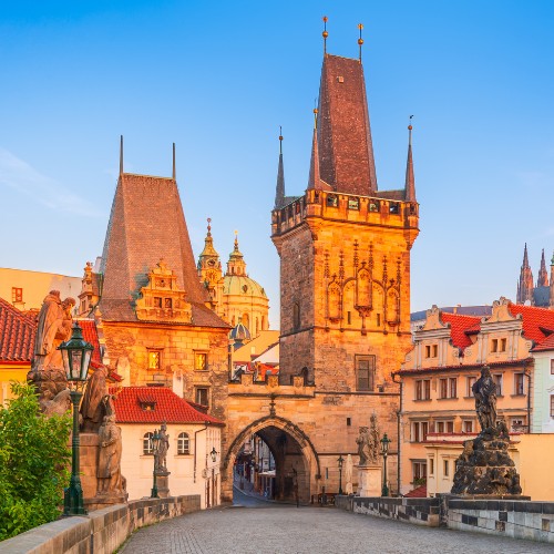 Charles Bridge in Prague at sunrise, featuring Gothic towers and statues. Warm light illuminates red-roofed buildings, creating a serene atmosphere.
