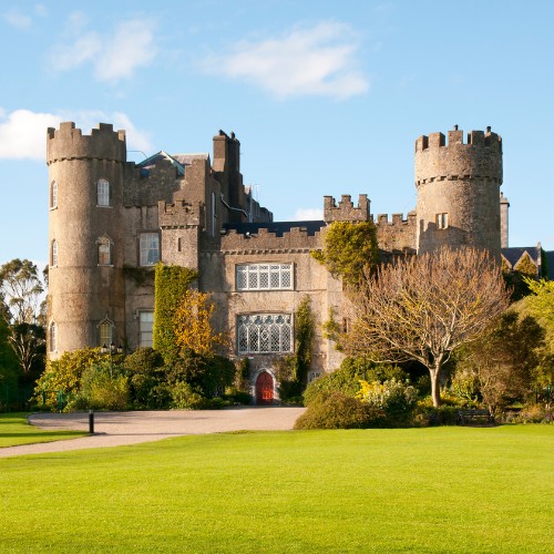 A historic stone castle with round towers stands amidst lush greenery under a blue sky. The scene conveys a sense of grandeur and tranquility.