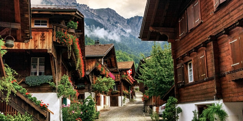 Charming alpine village with wooden chalets adorned with vibrant flowers. A cobblestone path leads to misty mountains, evoking tranquility and rustic beauty.