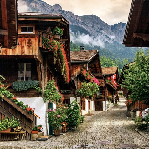 Charming alpine village with wooden chalets adorned with vibrant flowers. A cobblestone path leads to misty mountains, evoking tranquility and rustic beauty.