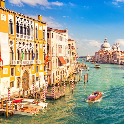 Majestic Venice canal scene with colorful historic buildings, gondolas, and boats on vibrant turquoise water under a clear blue sky with distant domes.