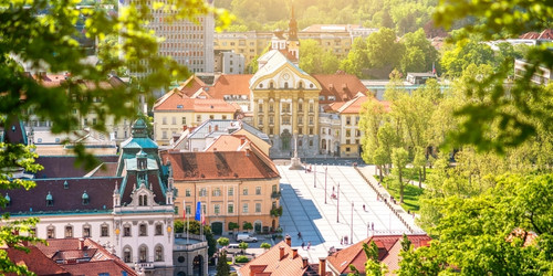 Aerial view of a European city with historic architecture, red-roofed buildings, and a tree-lined plaza under bright sunlight, evoking a serene ambiance.