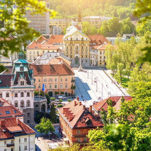Aerial view of a European city with historic architecture, red-roofed buildings, and a tree-lined plaza under bright sunlight, evoking a serene ambiance.