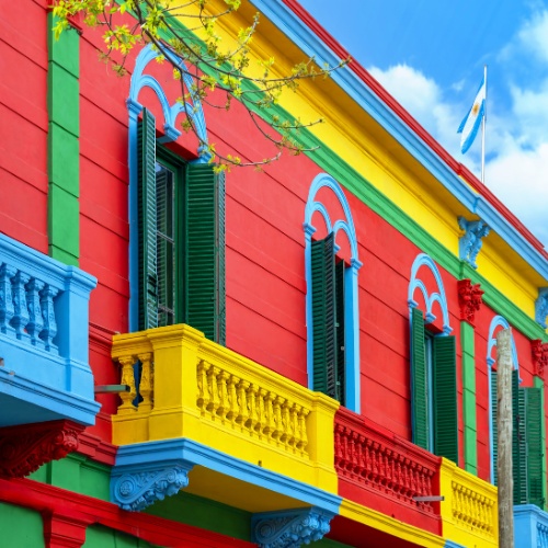 Colorful building facade with vibrant red, yellow, blue, and green accents, featuring balconies and an Argentine flag against a blue sky.