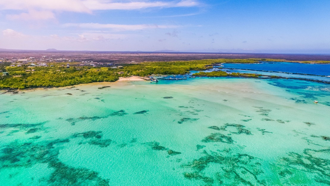 Aerial view of a pristine turquoise lagoon with coral reefs, bordered by lush green vegetation and a distant shoreline under a bright blue sky.