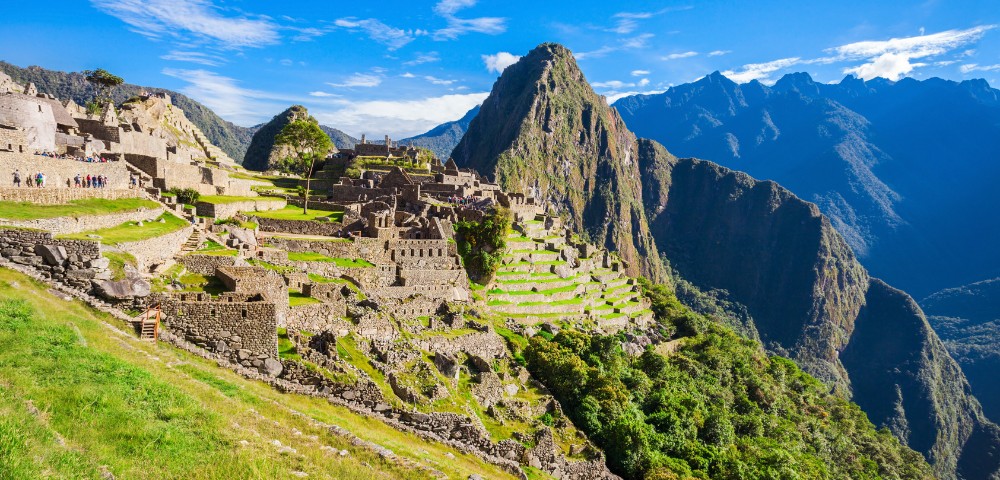 A breathtaking view of Machu Picchu with terraced ruins cascading down lush green slopes, surrounded by towering Andes mountains under a clear blue sky.