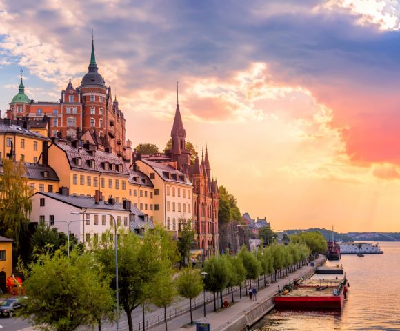 Sunset view of Stockholm's historic waterfront, featuring colorful buildings with spires, lush trees, and calm water reflecting vibrant sky hues.