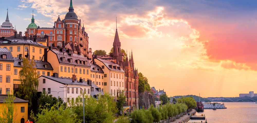 Sunset view of Stockholm's historic waterfront, featuring colorful buildings with spires, lush trees, and calm water reflecting vibrant sky hues.