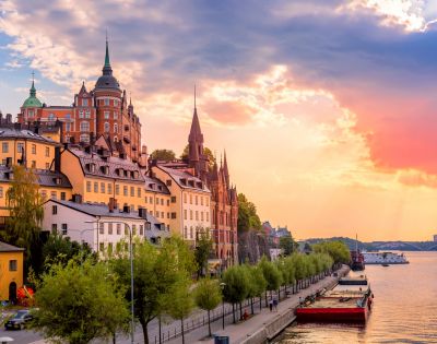 Sunset view of Stockholm's historic waterfront, featuring colorful buildings with spires, lush trees, and calm water reflecting vibrant sky hues.