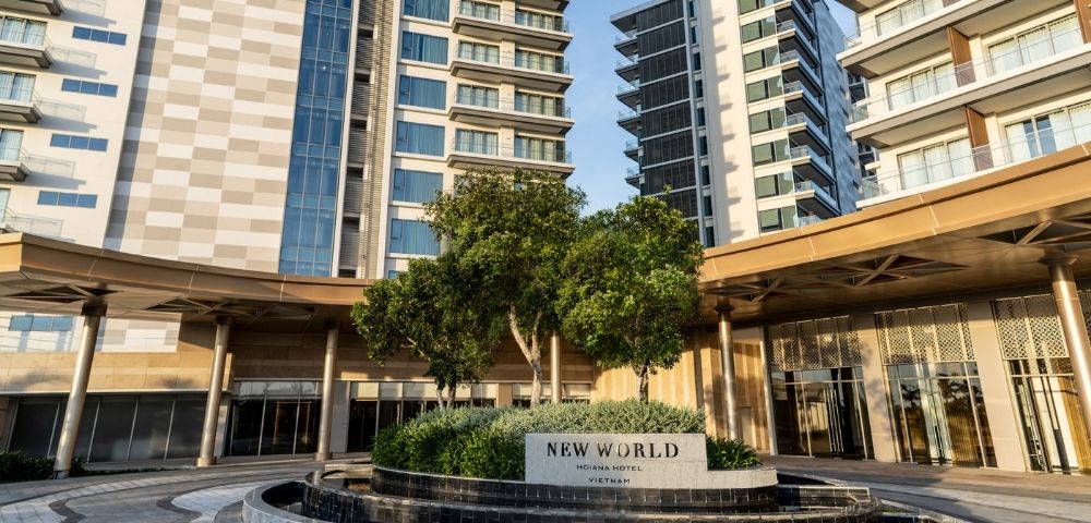 Entrance of New World Hoiana Hotel in Vietnam with modern high-rise buildings and landscaped greenery under a clear blue sky.