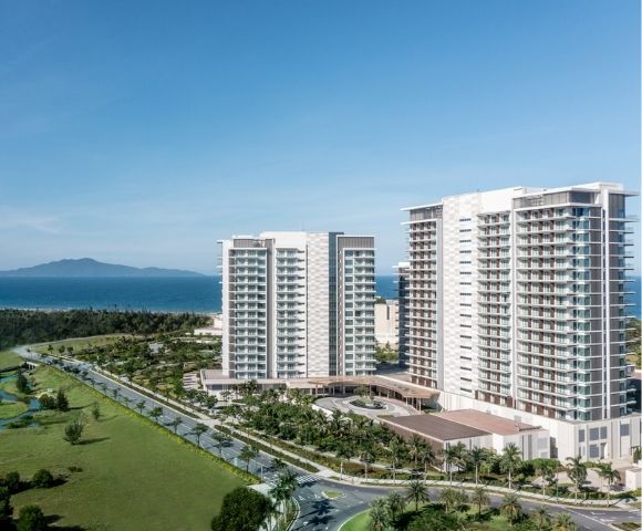 Modern high-rise buildings near a coastline with green fields, a winding road, and an island visible across the blue ocean under a clear sky.
