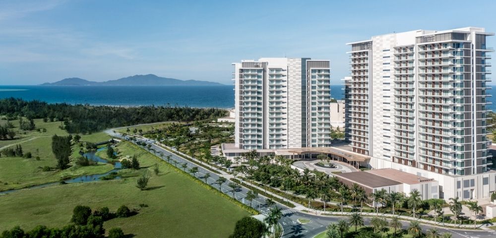 Modern high-rise buildings near a coastline with green fields, a winding road, and an island visible across the blue ocean under a clear sky.