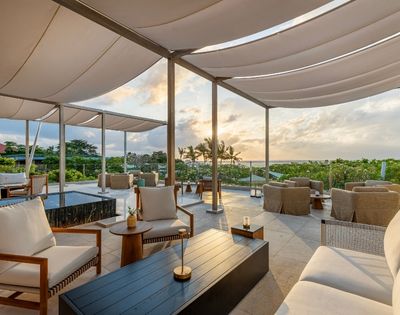 An outdoor lounge area with comfortable seating and a canopy roof overlooks a lush green landscape and the ocean at sunset.