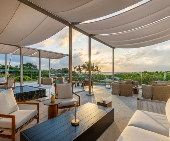 An outdoor lounge area with comfortable seating and a canopy roof overlooks a lush green landscape and the ocean at sunset.