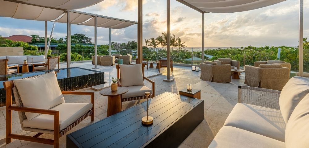 An outdoor lounge area with comfortable seating and a canopy roof overlooks a lush green landscape and the ocean at sunset.