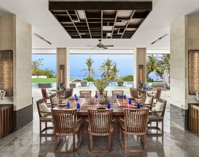 Open-air dining area with wooden table and chairs, overlooking palm trees, pool, and ocean under a clear blue sky.