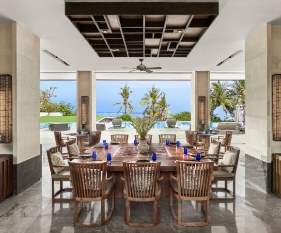 Open-air dining area with wooden table and chairs, overlooking palm trees, pool, and ocean under a clear blue sky.