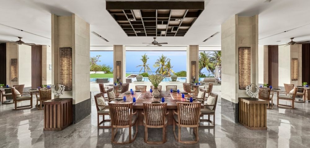 Open-air dining area with wooden table and chairs, overlooking palm trees, pool, and ocean under a clear blue sky.