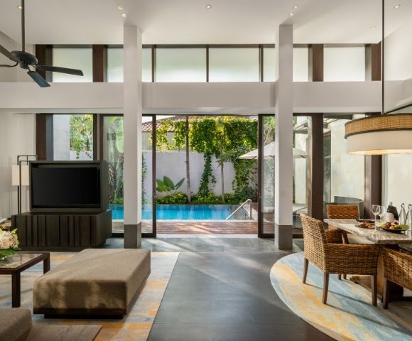 Modern open-plan living room and dining area with large glass doors opening to a pool and garden, featuring wicker chairs and neutral tones.