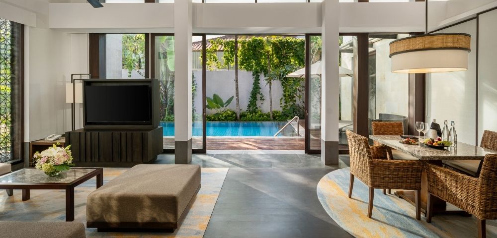 Modern open-plan living room and dining area with large glass doors opening to a pool and garden, featuring wicker chairs and neutral tones.