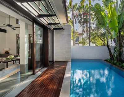 Modern outdoor pool area adjacent to a house with wooden deck, glass doors, ceiling fan, and tropical palm trees in the background.