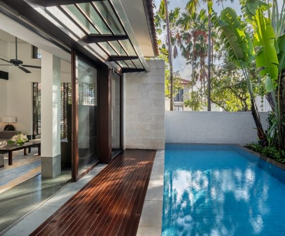 Modern outdoor pool area adjacent to a house with wooden deck, glass doors, ceiling fan, and tropical palm trees in the background.