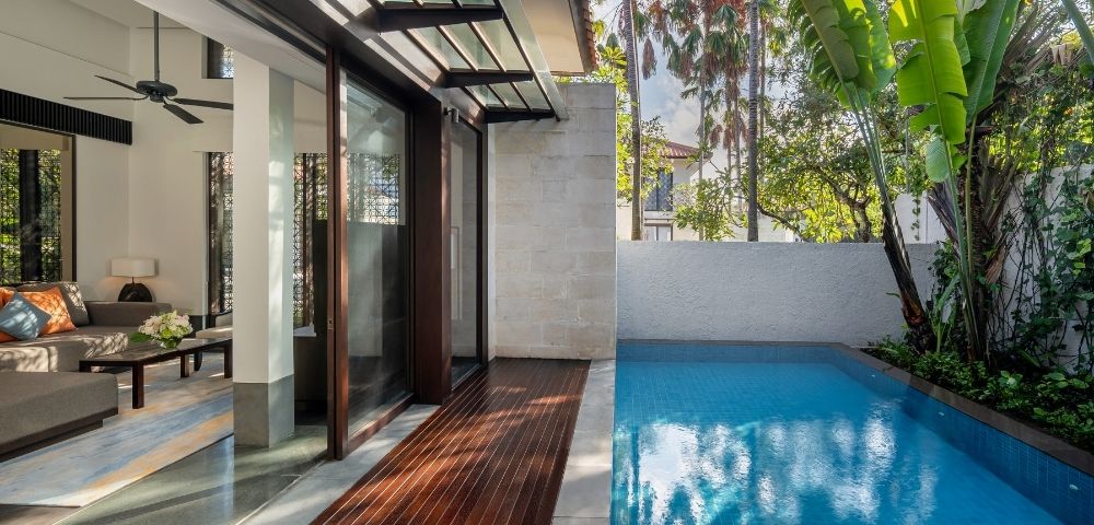 Modern outdoor pool area adjacent to a house with wooden deck, glass doors, ceiling fan, and tropical palm trees in the background.
