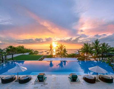 Infinity pool with lounge chairs and umbrellas overlooking palm trees and a vibrant sunset over the ocean.