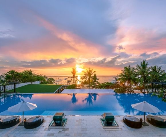Infinity pool with lounge chairs and umbrellas overlooking palm trees and a vibrant sunset over the ocean.