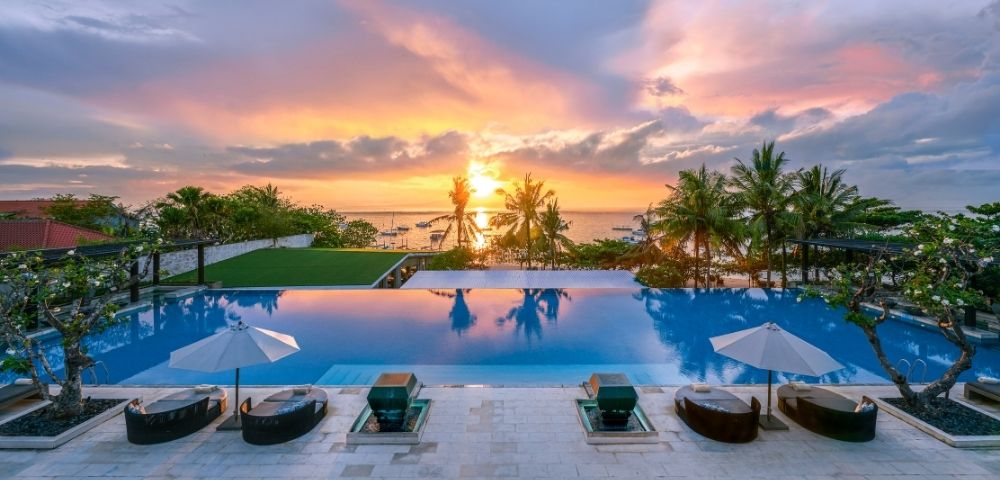 Infinity pool with lounge chairs and umbrellas overlooking palm trees and a vibrant sunset over the ocean.
